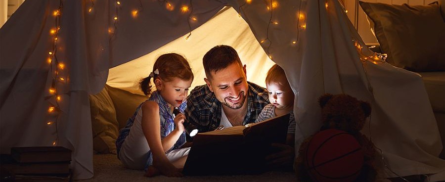 girls and father in a reading tent lit by fairy lights and flashlights.